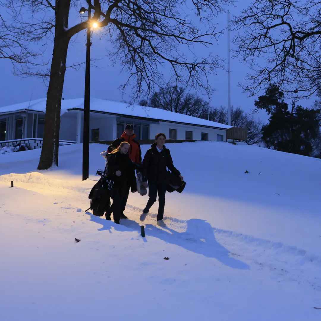 Three people walk through deep snow at dusk, carrying bags and golf clubs, under a streetlamp near a building and trees.