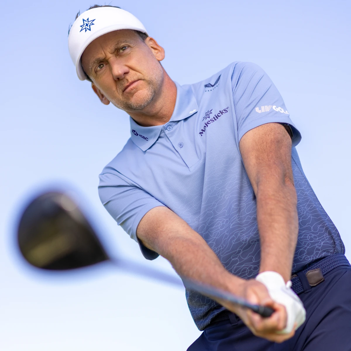 Golfer in a light blue shirt and cap, holding a golf club, looks focused against a clear sky background.