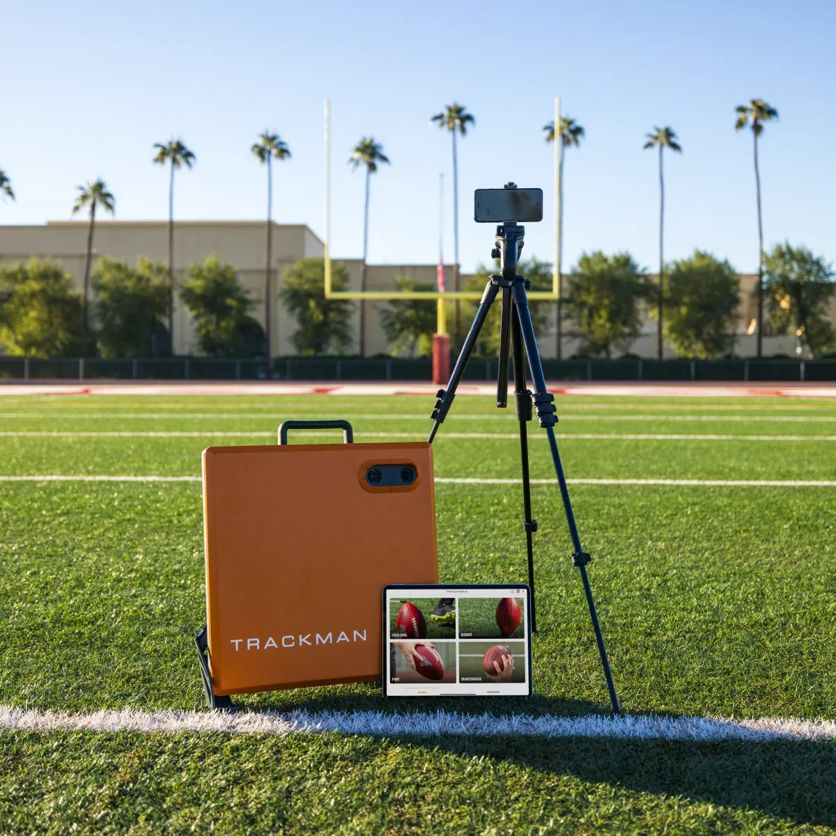 Tripod-mounted smartphone, orange TrackMan case and tablet displaying football shots on turf with goalposts and palm trees