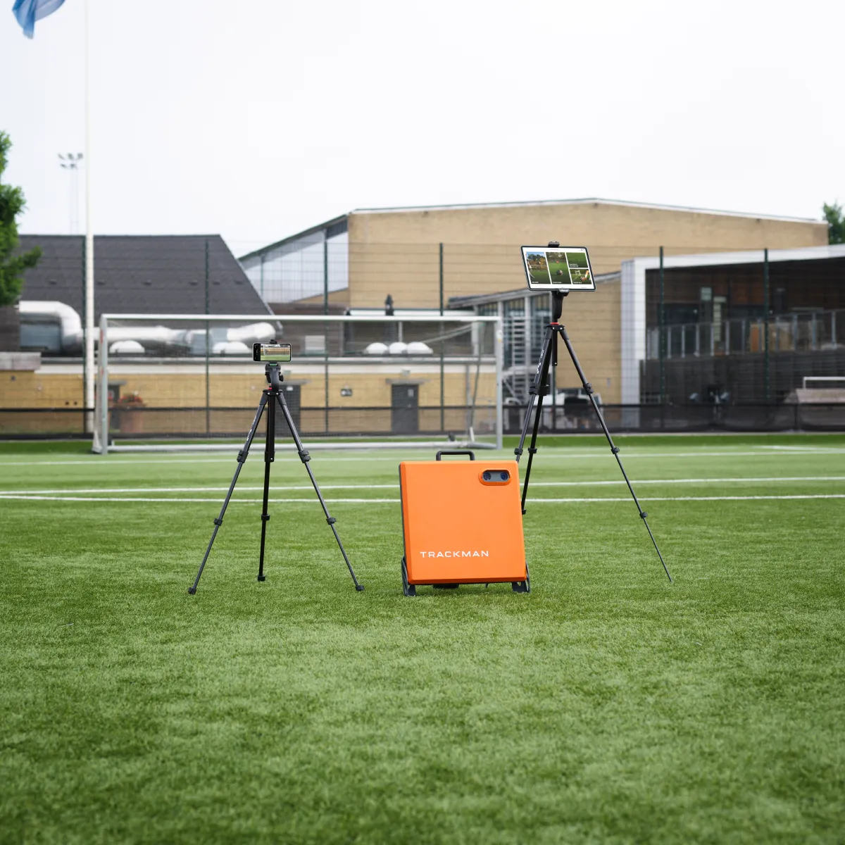 Orange TrackMan device and two tripods, one with a camera and another with a tablet, set up on a grassy sports field.