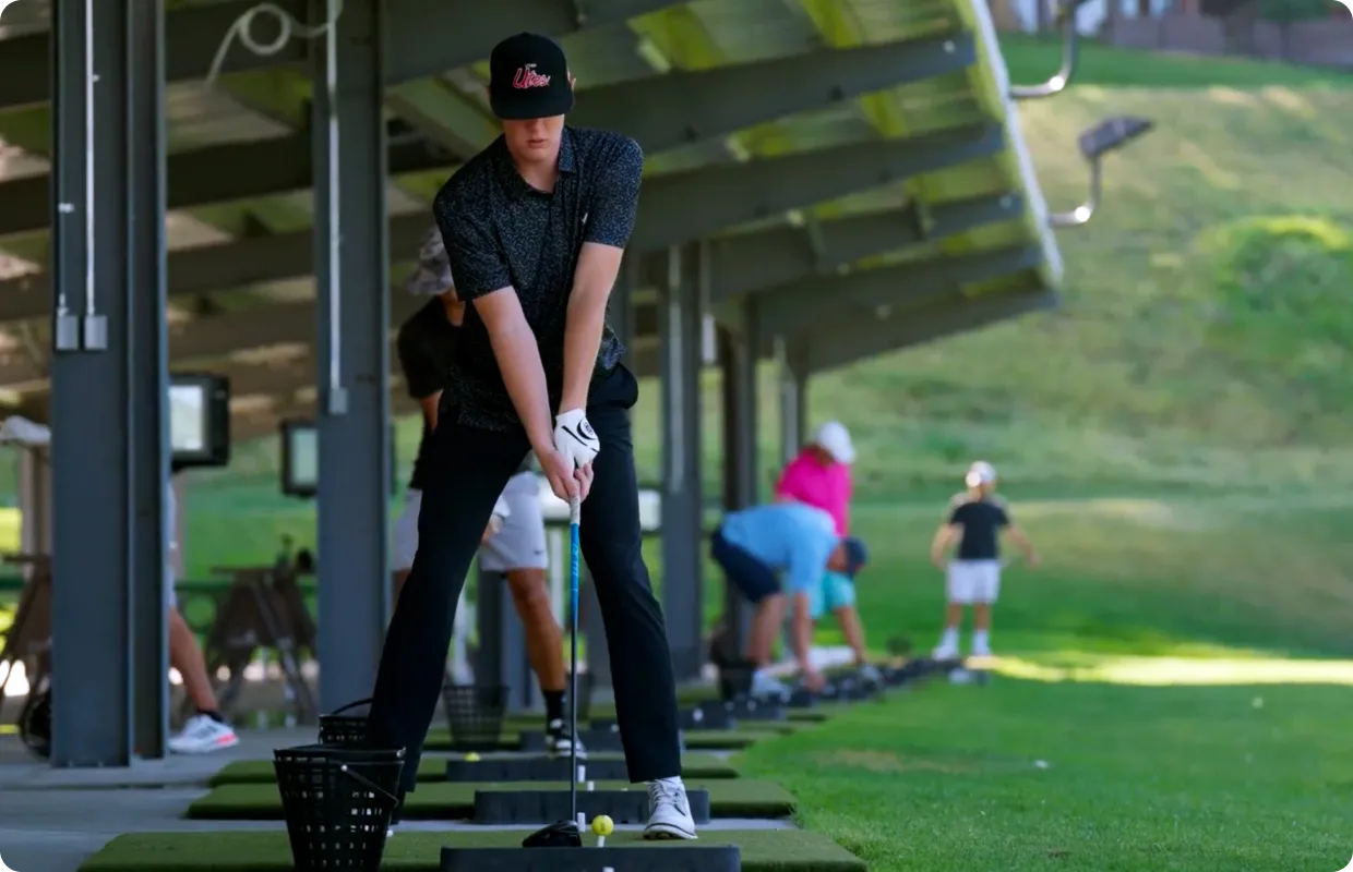 A person prepares to swing a golf club at a driving range, with others practicing in the background under a covered area.