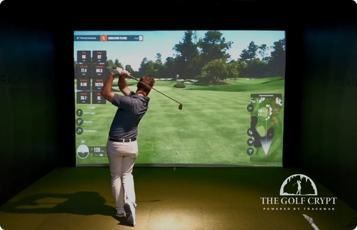 Man swinging golf club inside a simulator, facing a large projected golf course screen; "The Golf Crypt" logo visible at bottom right.