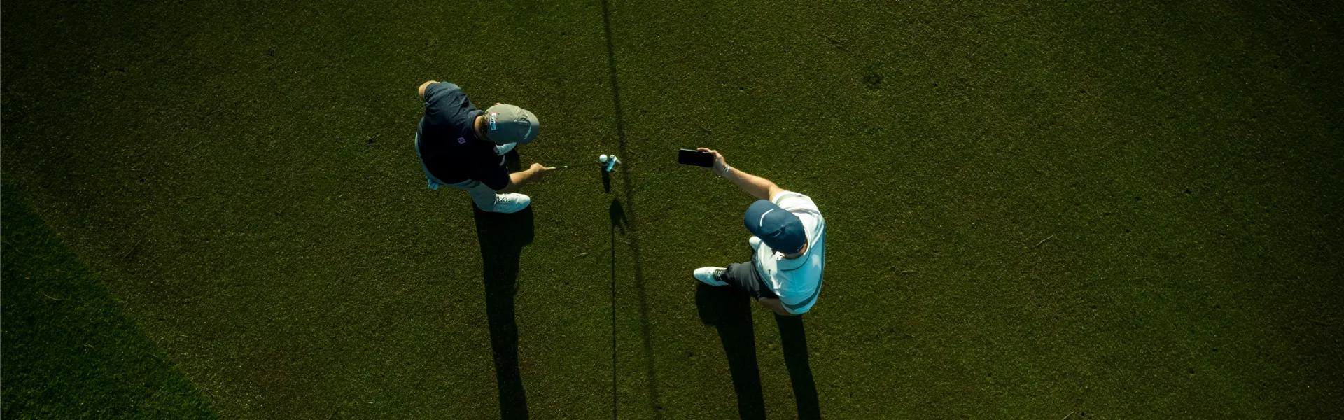 Two people on a golf course, seen from above, one takes a selfie while the other points at a golf ball on the grass.