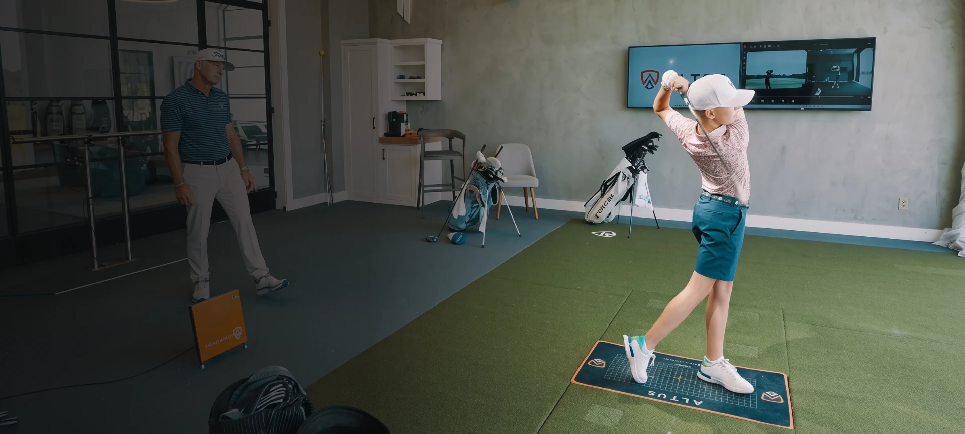 A young golfer practices a swing on an indoor green mat, with an instructor watching and golf equipment in the background.
