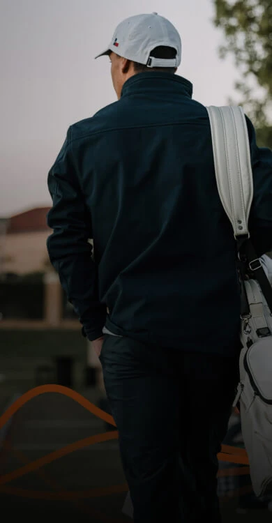 Man in a dark jacket and white cap carrying a white bag over his shoulder, walking outdoors at dusk.