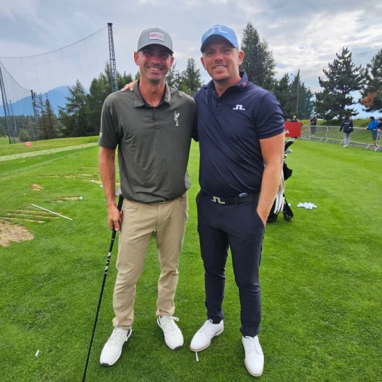 Two men smiling on a golf course, one in a grey polo and cap, the other in a navy outfit and cap, with golf clubs and trees in the background.