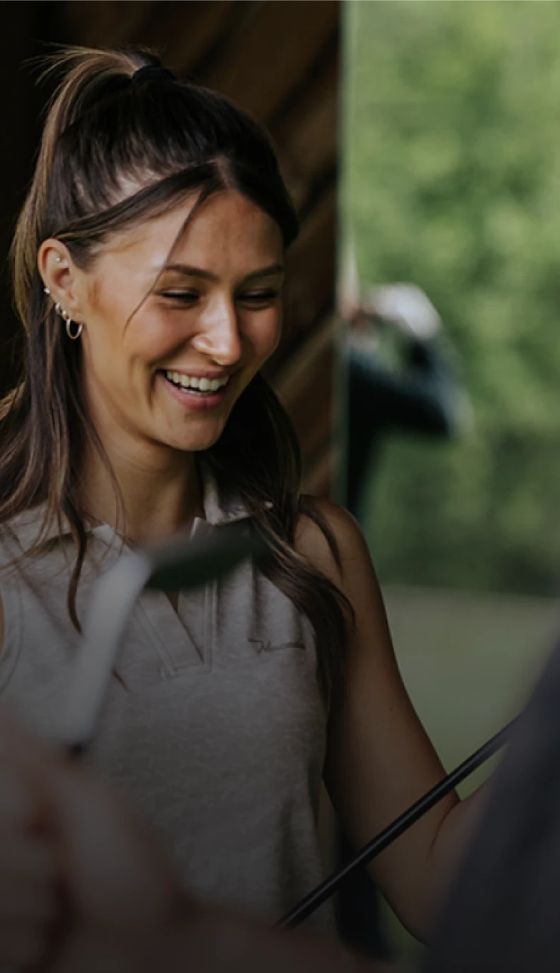 Smiling woman with dark hair holding a golf club, standing outdoors with a blurred background of greenery.