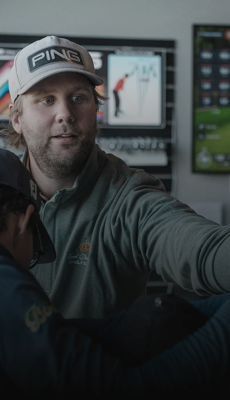 Man wearing a cap and jacket assists another person in a golfing store, with screens displaying golf-related content in the background.