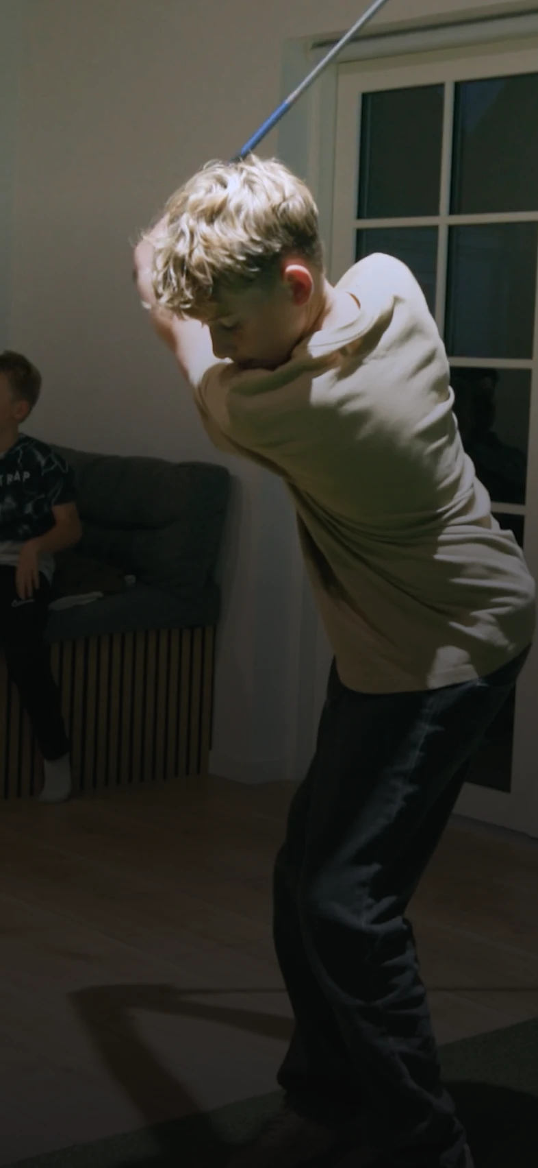 A person is swinging a game controller in a living room at night, while another person sits on a sofa in the background.