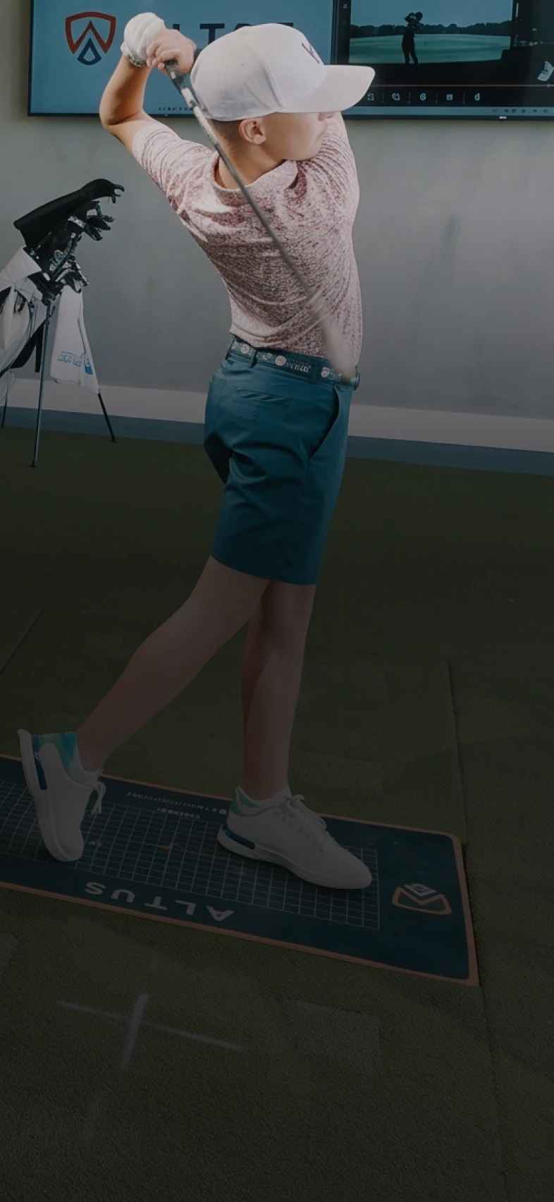 A golfer in a cap and casual sportswear completes a swing indoors, with a golf bag and screen in the background.