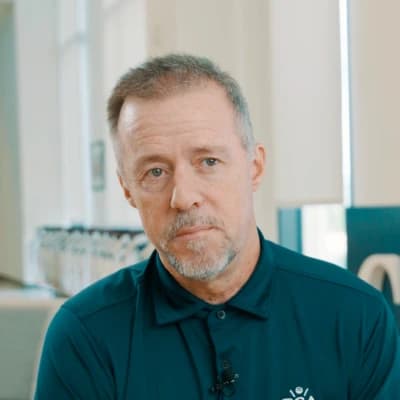 A man with short hair and a beard, wearing a dark teal polo shirt, looks directly at the camera in a well-lit room with large windows.