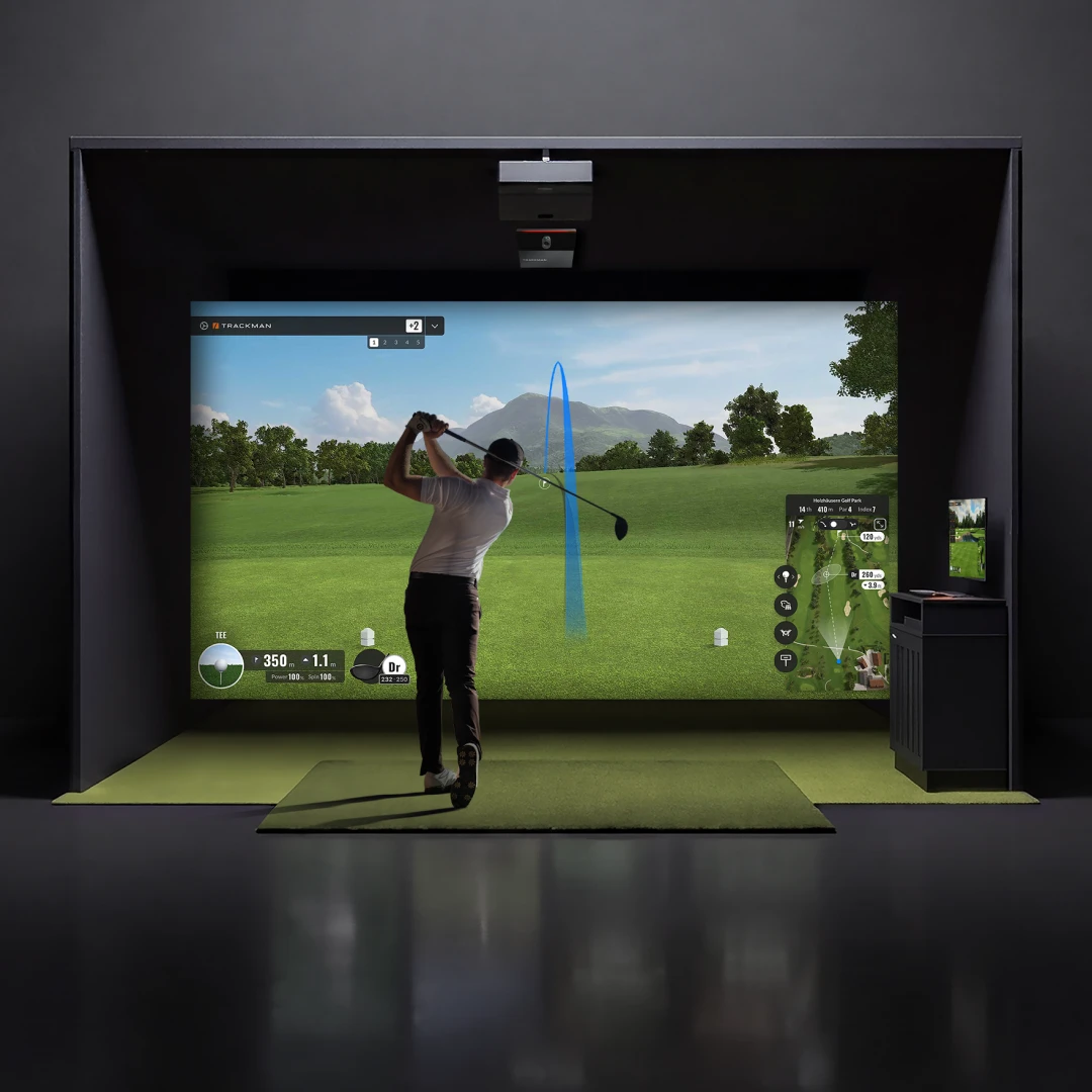 A person swings a golf club in an indoor golf simulator, facing a large screen displaying a virtual golf course.