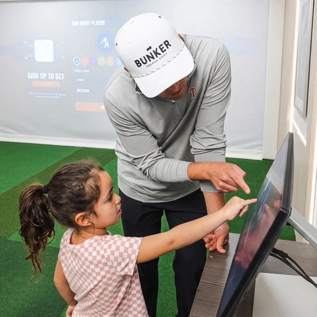A man and a young girl interact with a touchscreen in a room with a turf floor and digital displays. The man points, guiding the girl.