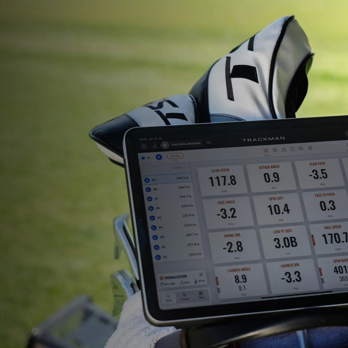 A tablet displaying golf swing metrics on a stand, with a golf bag and green field in the background.