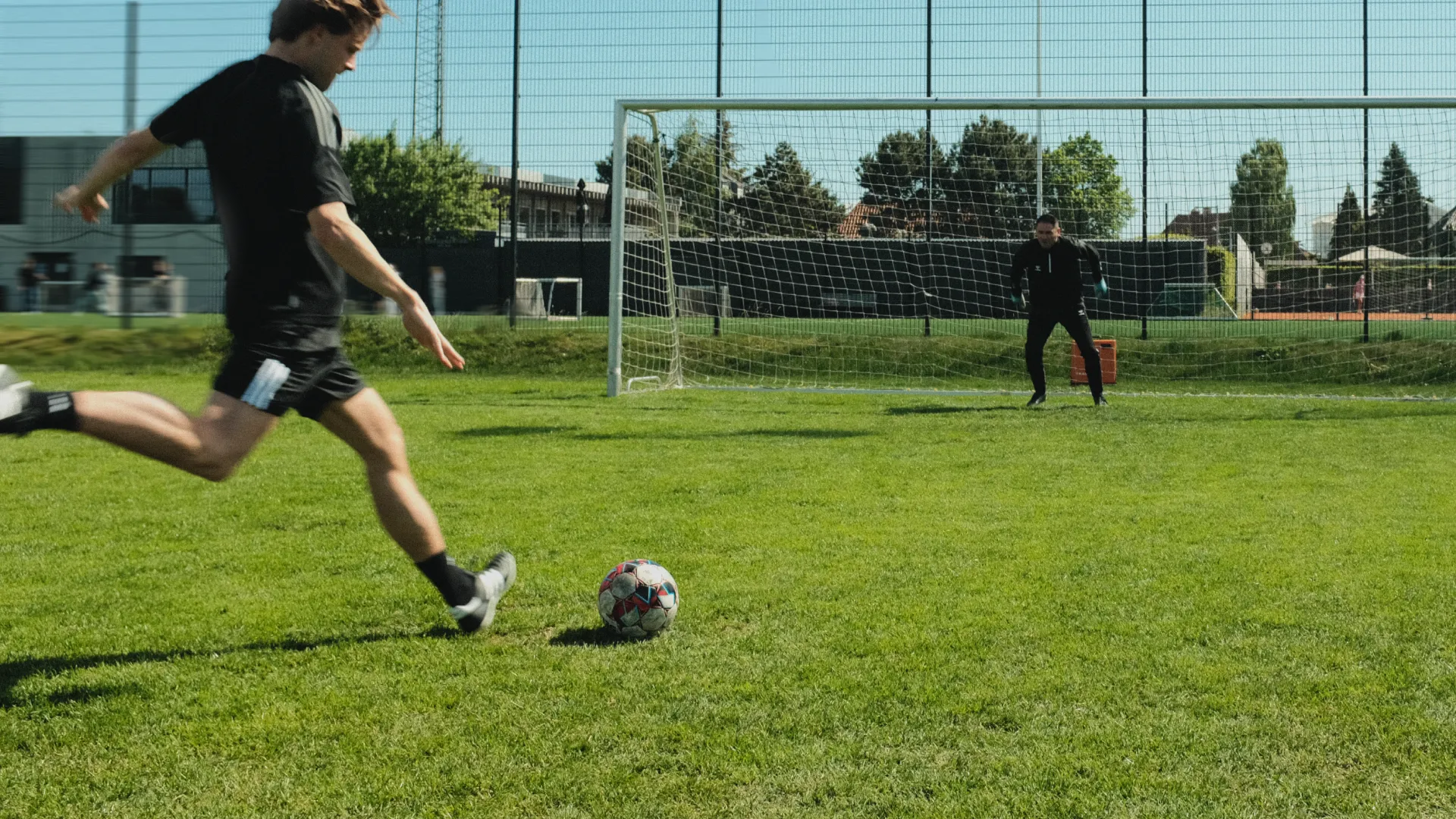 A soccer player in black prepares to kick a ball towards the goal, as the goalie gets ready to block on a sunny day.