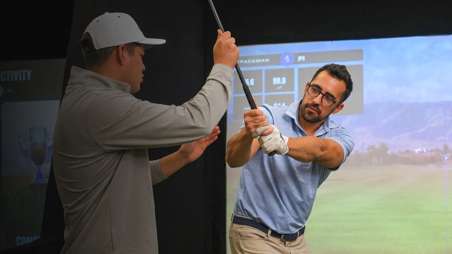 Man receiving golf swing instruction indoors, holding a club, while another person guides his form.
