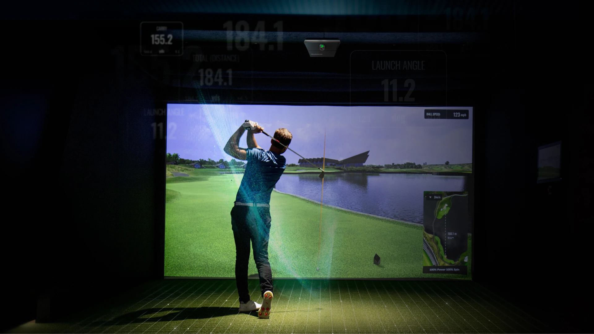 Person playing golf in an indoor simulator, swinging a club towards a projected course with water and greenery.