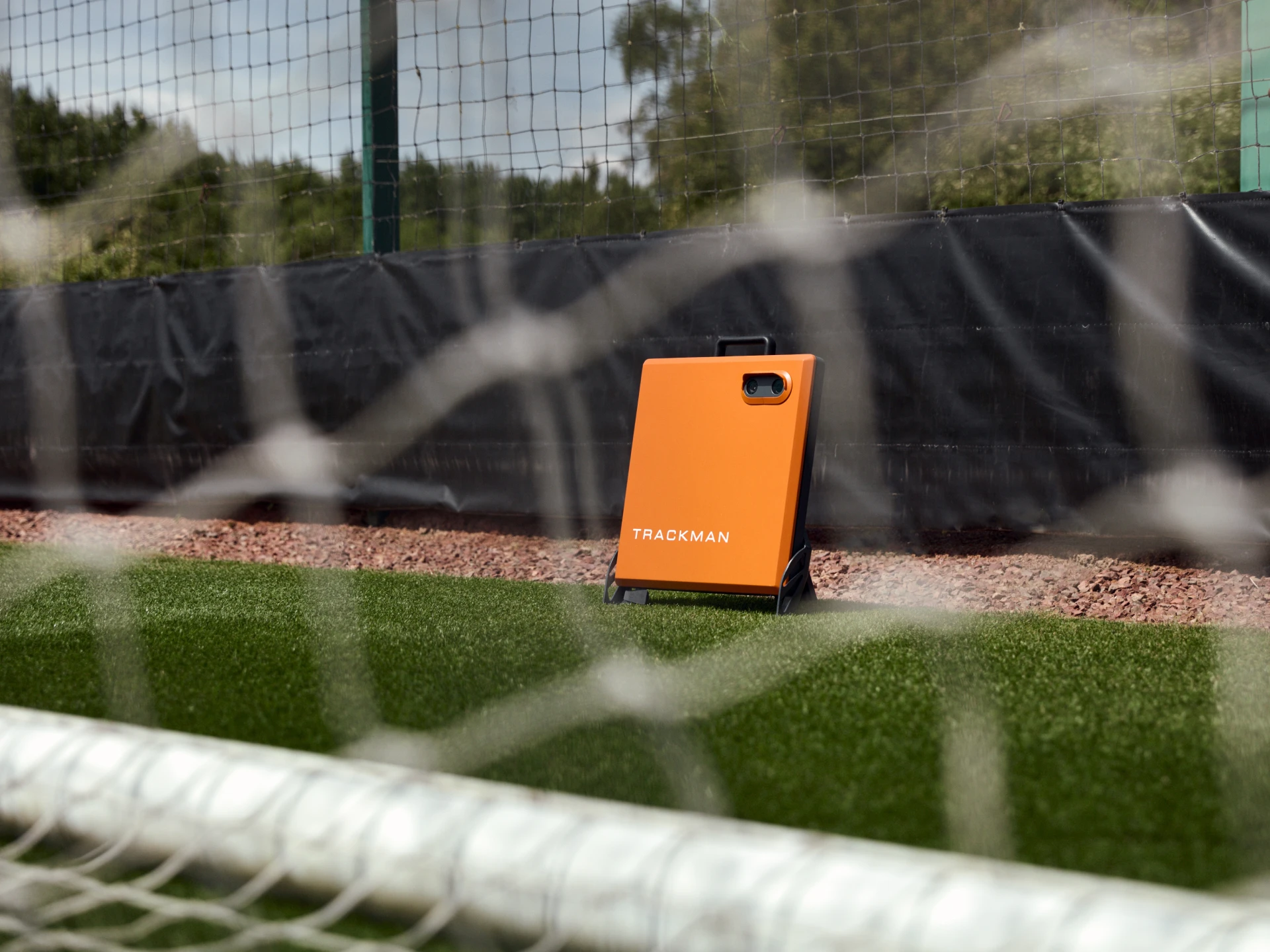 Orange Trackman device on grass by a soccer net, set against a black fence and trees in the background.
