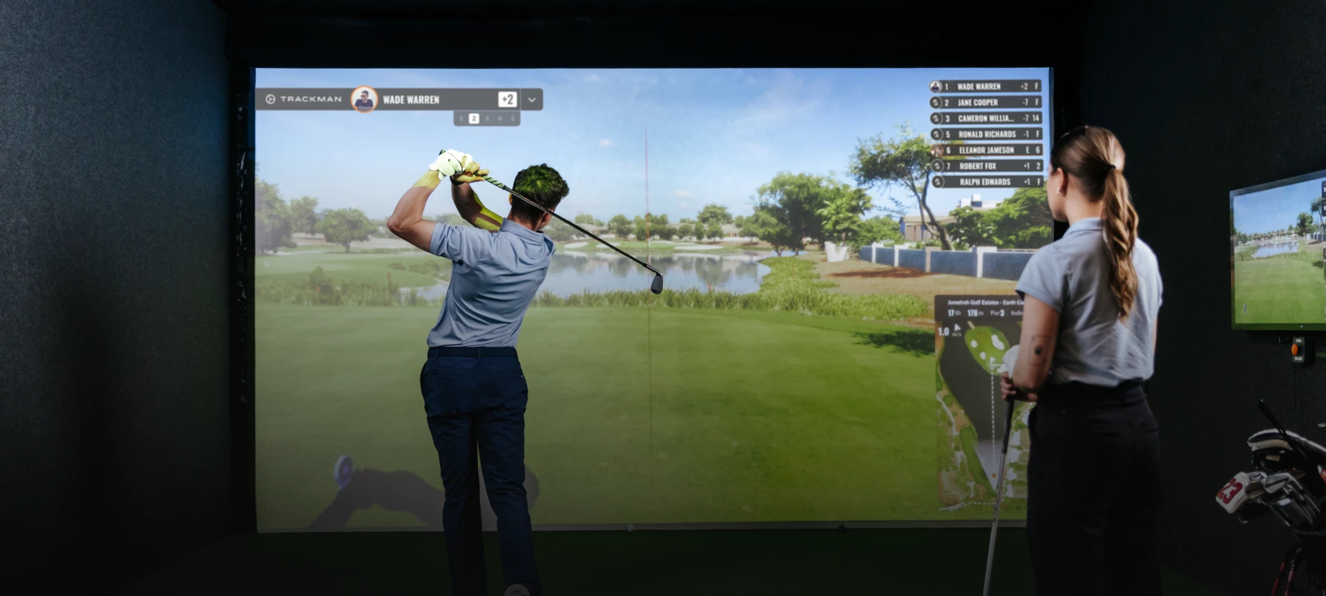 A man swings a golf club in front of a virtual golf simulator screen, with a woman observing. Scores are displayed on the screen.