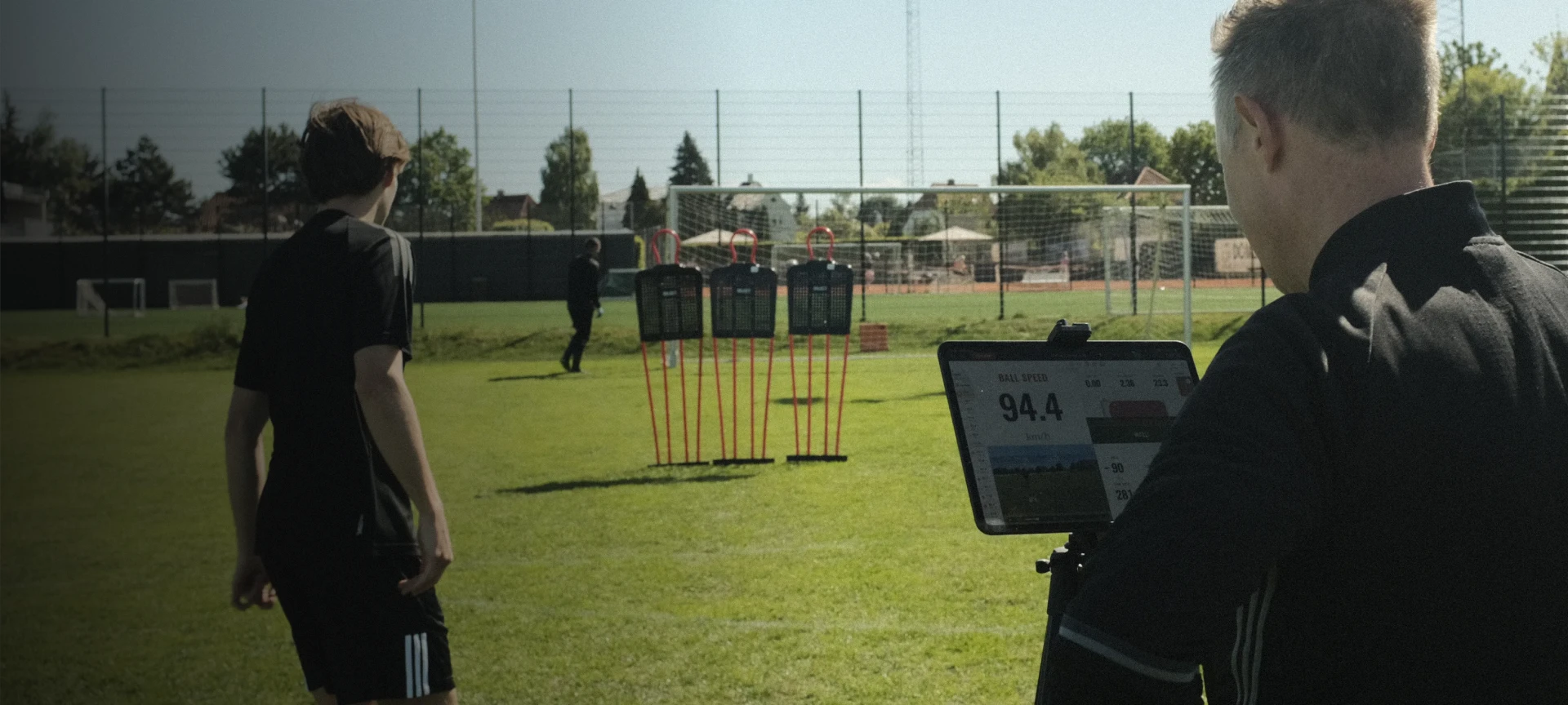 Coaches on a soccer field use a tablet to analyze a player's speed as they run towards agility poles near a goalpost.