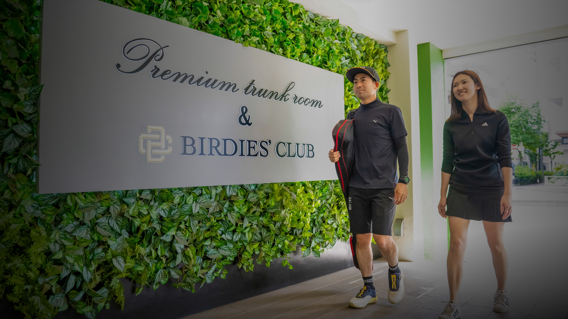 Two people in sports attire walk past a sign reading "Premium trunk room & Birdies' Club" on a leafy wall.