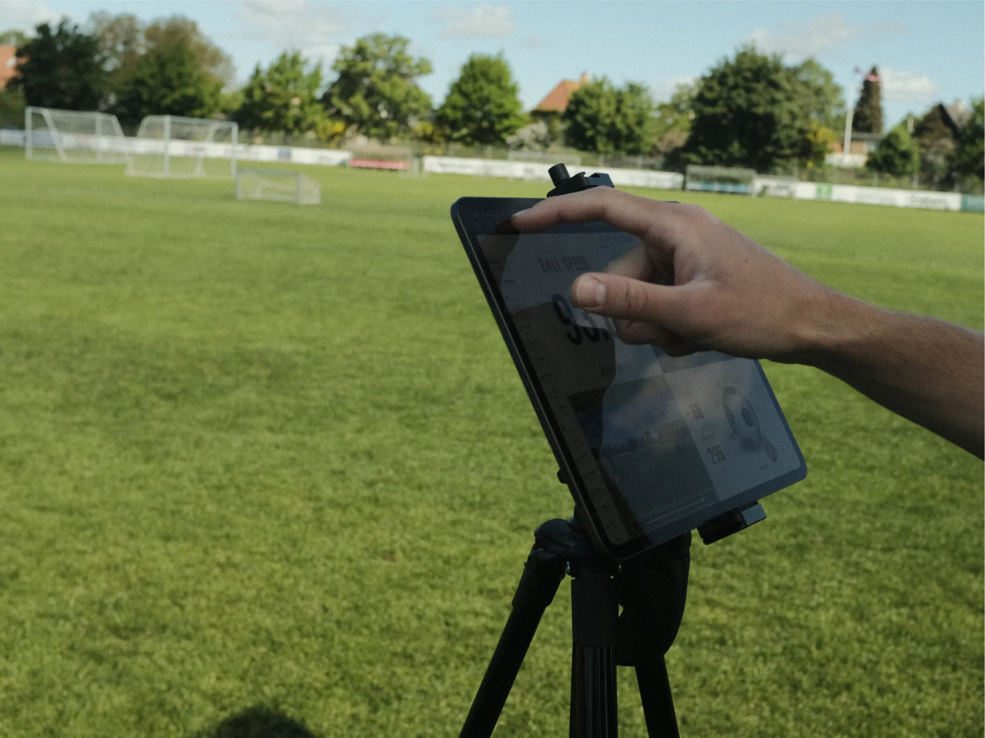 A hand interacts with a tablet on a tripod on a sports field, displaying data like ball speed. Soccer goals are visible in the background.