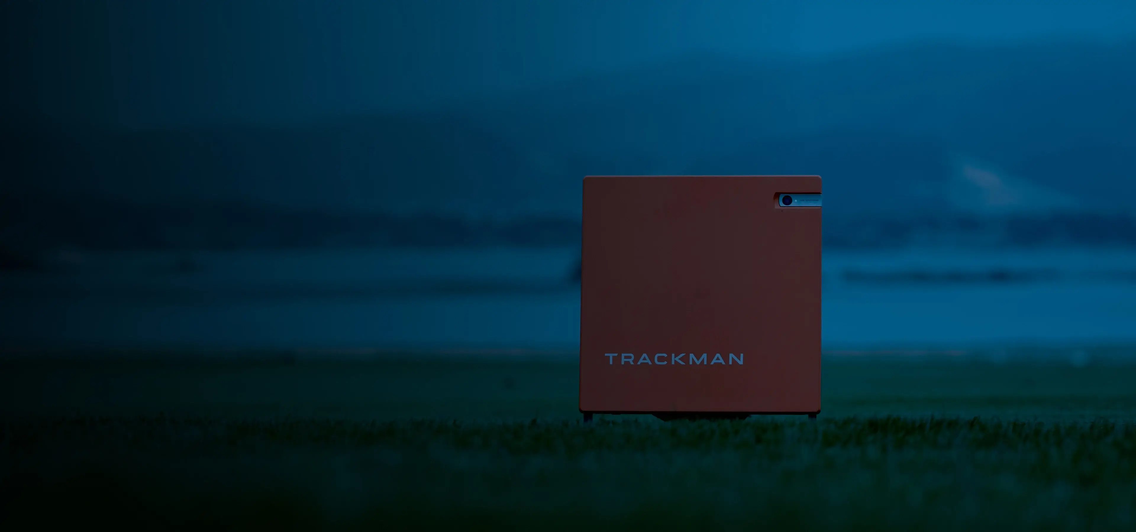 A red TrackMan device on grass at dusk, with a blurred mountainous background under a dark blue sky.