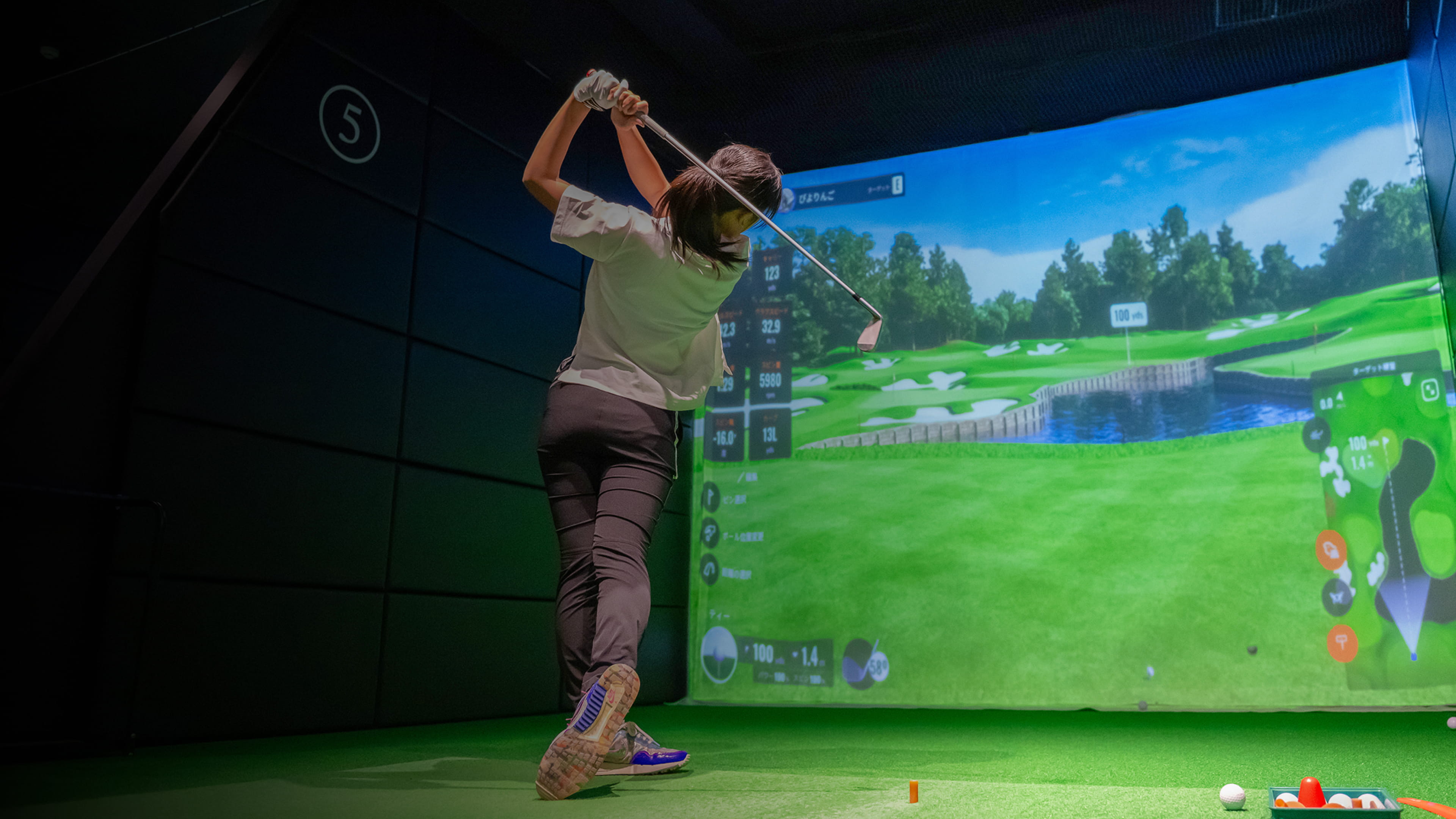 バーチャルコースが表示された屋内ゴルフシミュレーターのレーン5でスイングする人物。a