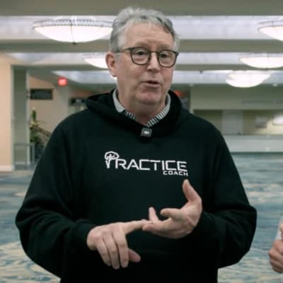 Person in glasses wearing a "Practice Coach" hoodie, speaking with gestures in a brightly lit indoor setting.