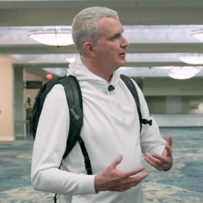Man in a white sweatshirt and backpack gesturing while talking in a brightly lit room with chandeliers and carpeted floor.