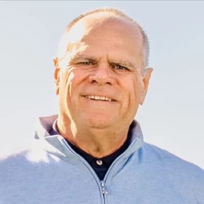 Smiling man in a light blue zip-up jacket against a clear sky background.