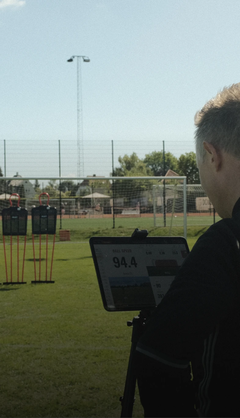 Person monitoring ball speed on a laptop, showing 94.4 km/h, with training mannequins and goalposts on a grassy field.