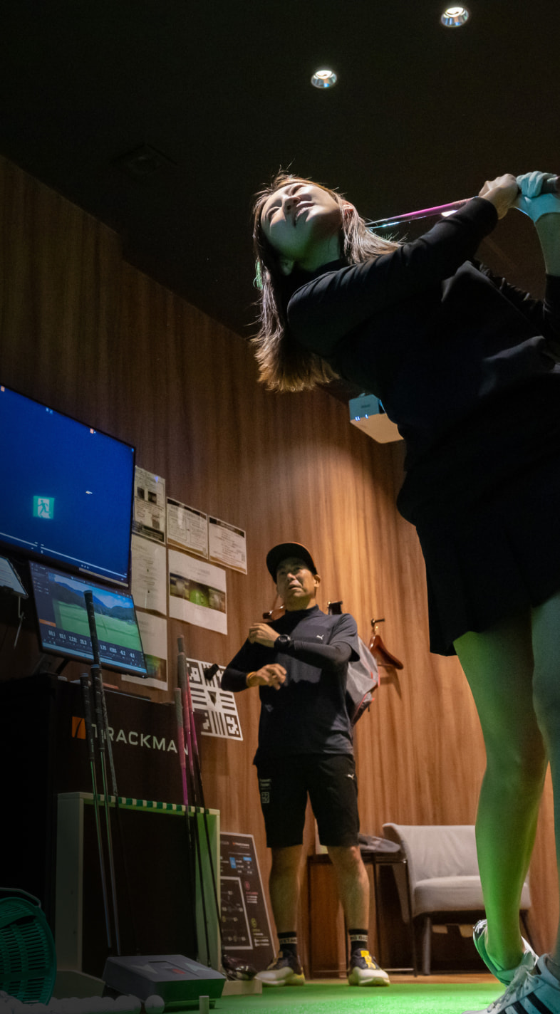 A woman swings a golf club indoors while a man watches. The room has a wooden wall, a monitor, and golf equipment.