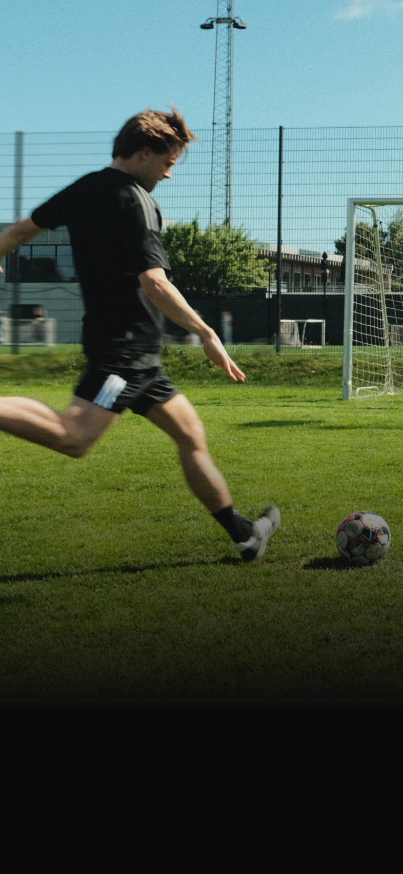 A soccer player in black kit kicks a ball on a grassy field, with a goalpost and fence in the background on a sunny day.